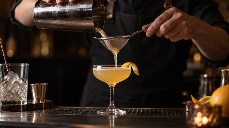 A bartender's hands fine-straining a pale sour into a coupe glass with a citrus twist on a dark bar counter