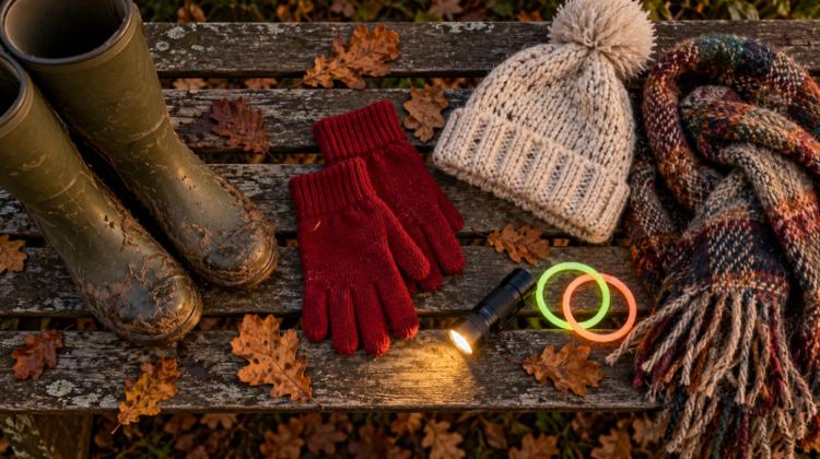 Muddy wellies, knitted gloves, a woolly hat, a scarf and a torch laid out on a wooden bench with autumn leaves