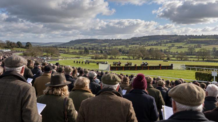A race day crowd in tweed coats and flat caps watching horses jump a fence on a green course below Cotswold hills