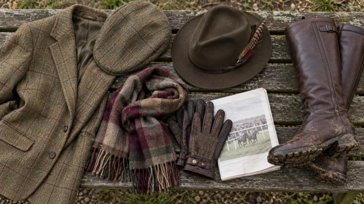 A tweed jacket, flat cap, feathered felt hat, scarf, leather gloves, a racecard and muddy knee-high boots laid on a wooden bench