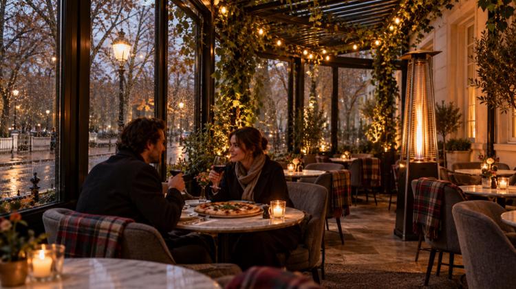 A couple sharing pizza and red wine in a glass conservatory with a patio heater, blankets and string lights on a rainy autumn evening
