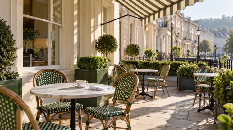 Green rattan bistro chairs and marble tables under a striped awning outside a cream stucco Regency terrace on a sunny morning