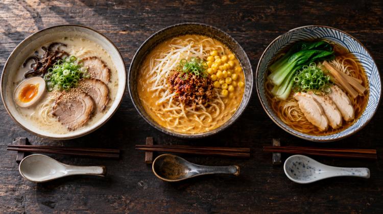 Three bowls of ramen on a dark wooden table: a cloudy pork tonkotsu, a golden miso with corn and a clear soy broth with chicken