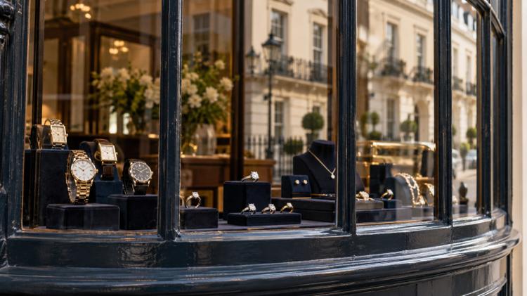 Watches and rings on dark velvet stands in the bow window of a traditional jeweller, with a cream Regency terrace reflected in the glass