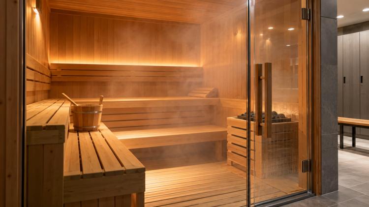 An empty cedar sauna with wooden benches, a bucket and ladle, and a glass door onto a changing room