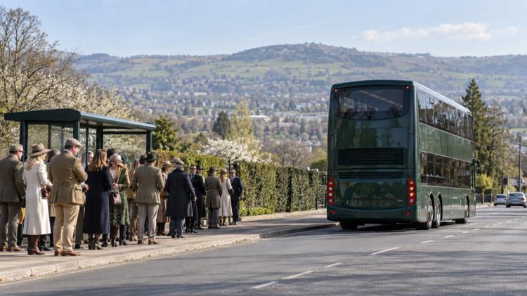 Racegoers in tweed queuing at a bus stop as a green double-decker pulls away on the edge of Cheltenham, with hills behind