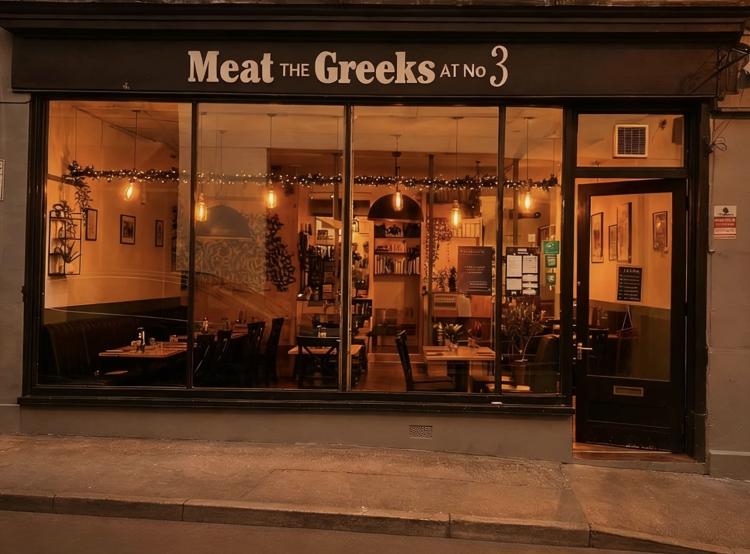 The glass shopfront of Meat the Greeks at No3 on Bennington Street, Cheltenham, lit up at dusk