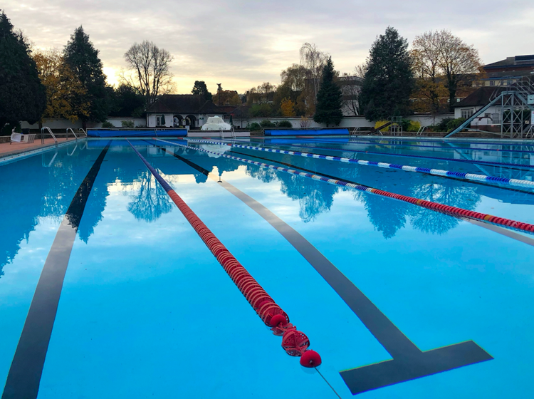 Lane ropes across the still 50m pool at Cheltenham Lido at dusk, with a slide tower and autumn trees behind