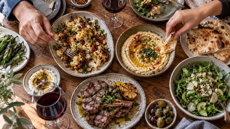 Sharing plates of roasted cauliflower, hummus, grilled lamb and salad on a wooden table with glasses of red wine