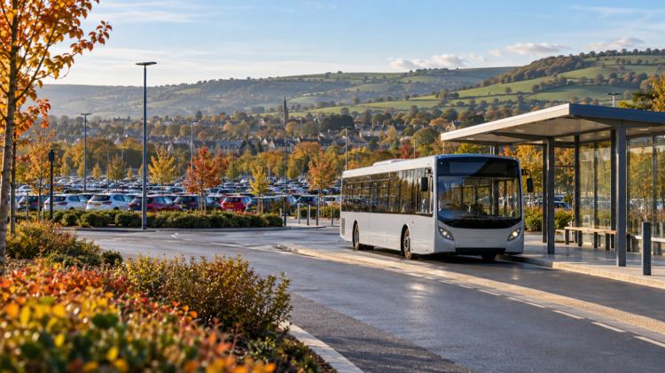 A single-decker bus pulling into a covered stop beside a large car park on the edge of Cheltenham, with green hills behind
