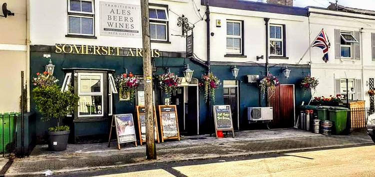 The dark blue front of the Somerset Arms on Moorend Street, Cheltenham, with hanging baskets and a Union flag