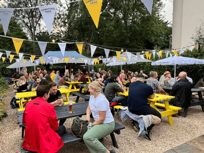 Drinkers at yellow picnic tables under bunting in the beer garden at Sandford Park Alehouse, Cheltenham